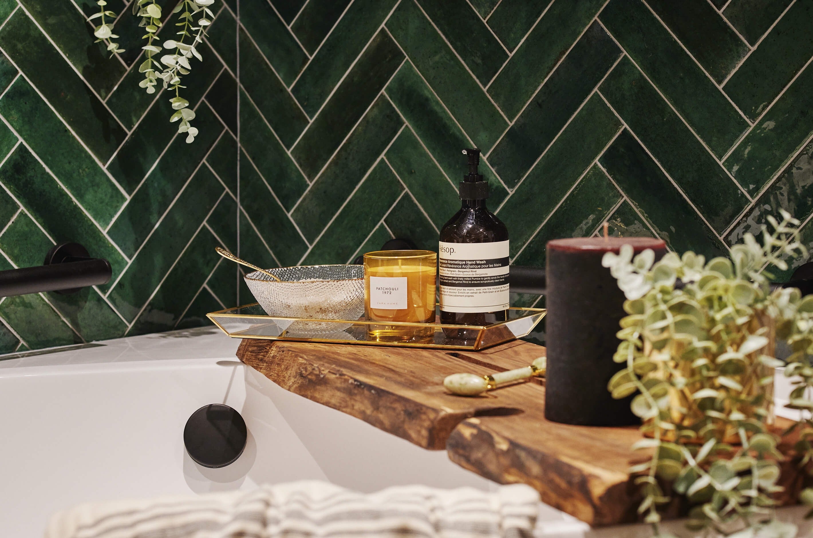 Wooden bath board resting on a corner bath against a backdrop of rich green tiles in a herringbone pattern and matt black fixtures.