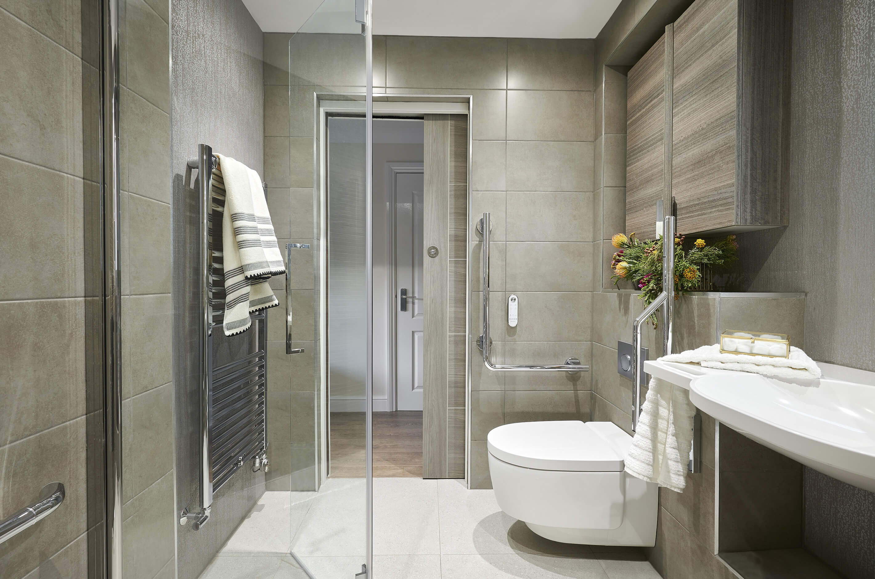 Natural-colour level access shower room with gold accents, with wash and dry toilet, grab rails and sliding door looking into hallway.